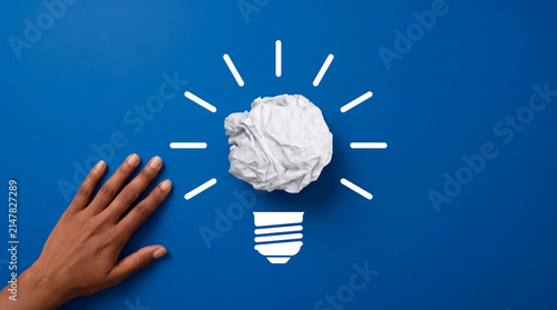 Creative business idea concept with a crumpled paper lightbulb and a human hand on a solid blue background