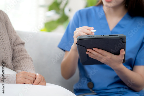 Asian woman caregiver using digital tablet for senior patient consultation, professional nurse checking health record in living room at home, elderly care assessment with checkup, health care support.