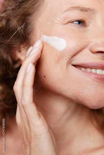 Obraz na plátně Authentic portrait of a mature woman applying cream to her cheek, natural skin with no retouching, highlighting skincare routine and selfcare for glowing complexion