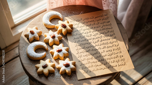 Cookies arranged beside a parchment sheet featuring elegant Arabic calligraphy sit upon a round wooden table by the window.