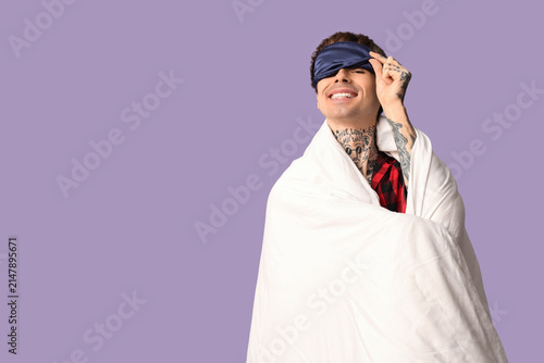 Handsome young man with blanket and sleeping mask on lilac background