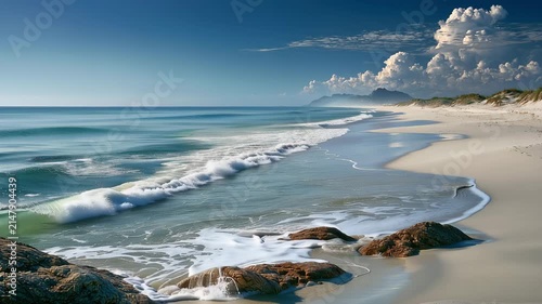 Crystal clear turquoise ocean waves washing onto pristine white sandy beach under blue sky with peaceful tropical coastline