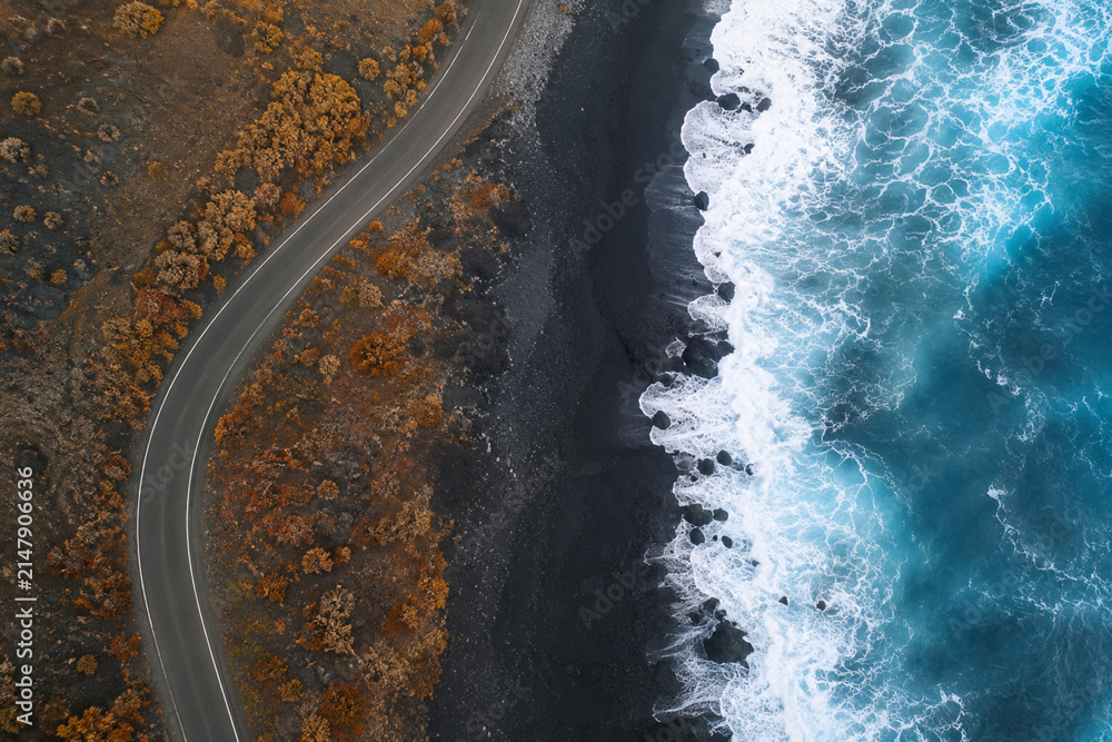 Obraz premium Aerial perspective of a winding road along a volcanic black sand beach with crashing blue ocean waves.