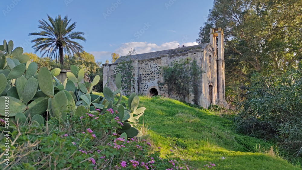 Naklejka premium Scenic view of the historic Acendu Church (Agios Georgios) in Lefke, set on a grassy hill surrounded by lush Mediterranean vegetation, palm trees, and prickly pear cacti.