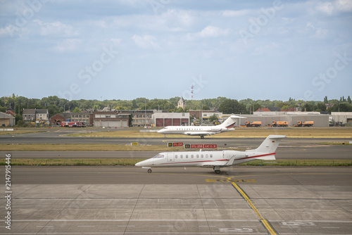 Two private business jets taxiing on parallel taxiways at an international airport with airport buildings and ground service vehicles in the background.