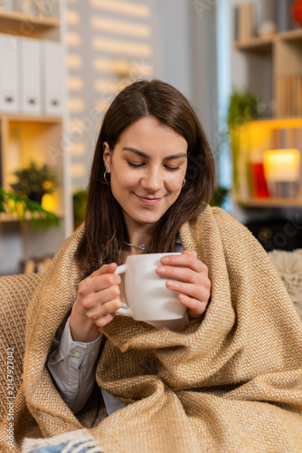 Quadro su tela Young woman on home sofa warms up with hot drink under blanket enjoying quiet rest after work in living room