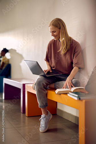 Happy college student surfing the net on laptop while learning in hallway.