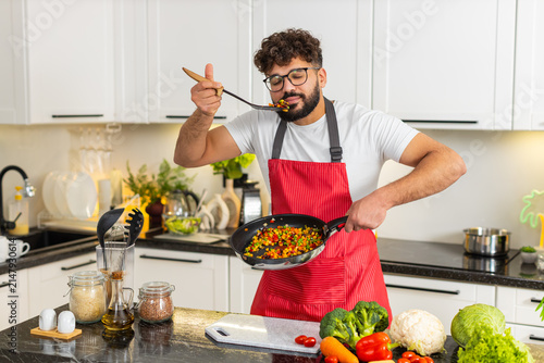 Canvas Print Arabian man stirs vegetables in pan and tastes spoonful in kitchen preparing healthy dinner for diet nutrition and fitness goals