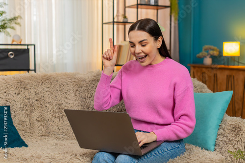 Young excited woman at home working on laptop actively seeking solution to issue, suddenly shouting Eureka. Caucasian girl on sofa happy and excited, successfully finding answer with joyful expression