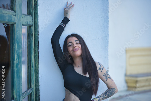 Young Hispanic woman, brunette, wearing a black top and jeans, heavily tattooed, smiling and happy, leaning against the frame of a glass door of an old building in the city.