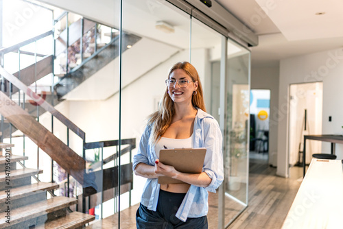 Office corridor portrait of a business woman. Corporate management and administration, Smiling female employee holding clipboard and pen walking in modern office hallway near stairs.
