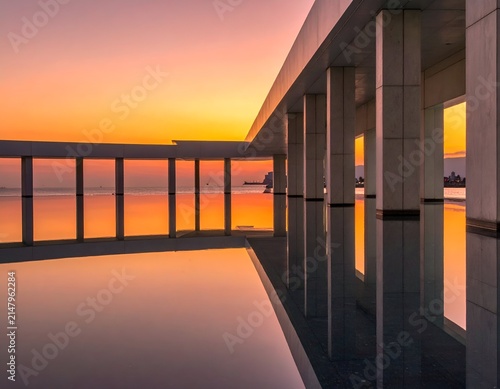 Modern building overlooking water at sunset, reflecting colorful sky and architecture