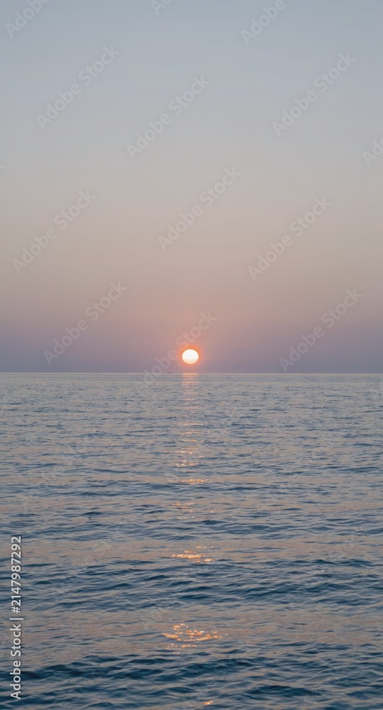 Obraz premium Sun setting over calm ocean horizon with soft pastel sky and reflective water surface, tranquil seascape at dusk conveying peaceful evening mood.