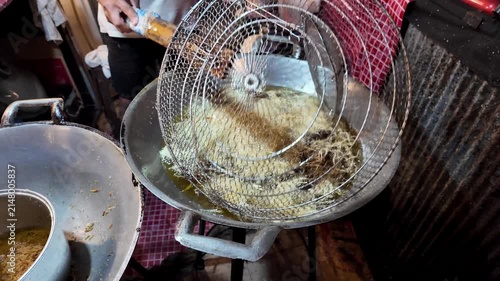 Close-up of freshly poured traditional Thai funnel cake frying in bubbling hot oil inside a large wok at a busy night market. Authentic Asian street food dessert being deep fried with visible oil bubb