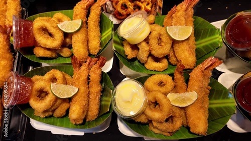 Assorted deep fried seafood platters served with crispy breaded shrimp, squid rings, dipping sauces and lemon wedges at a Thai street food market. Delicious seafood takeaway meals presented on banana 