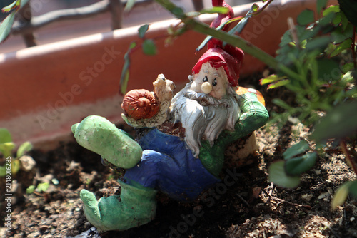 Piccola statuina decorativa di nano da giardino seduto su una poltrona verde in miniatura all'interno di una fioriera con terriccio e piante sul balcone