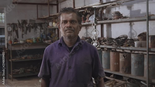 A middle aged Indian skilled, confident male fabricator worker in a uniform, standing and facing or looking at the camera in a fabrication workshop surrounded by the tools, equipment, and raw material