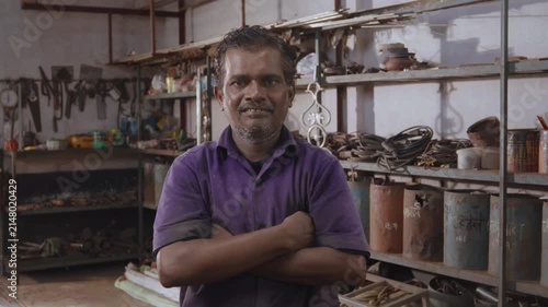 Middle-aged Indian, happy, confident skilled male fabricator wearing a uniform, standing with crossed arms, looking at the camera, smiling in a fabrication workshop surrounded by tools and equipment
