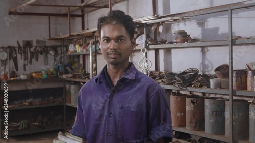 A young Indian skilled, confident, and happy male fabricator worker in a uniform, standing and facing or looking at the camera in a fabrication workshop surrounded by the tools, equipment, and raw mat