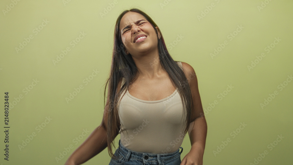 Obraz premium Young woman with long dark hair wearing beige tank top and high waist jeans rubbing neck and wincing in green studio setting; pain.