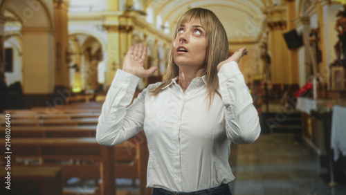 Woman raising hands and palms near ears while looking upward inside a building nave with pews and altar visible; prayerful contemplation.
