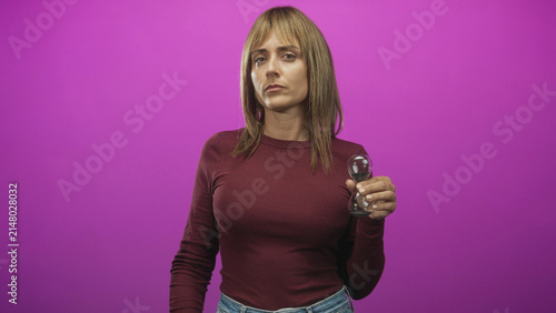 Fotografie Woman points finger while holding hourglass in studio purple backdrop maroon sweater; time pressure urgency