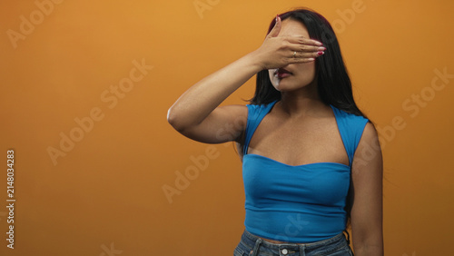 Photography Woman covering eyes with hand, palm over face, wearing blue strap top and jeans, long brunette hair visible against orange studio wall; shyness