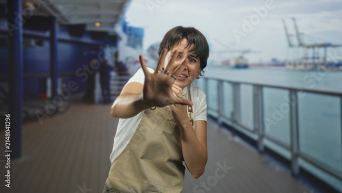 Fotografie Woman in apron shields face with outstretched hand on ship deck railing, wincing and gripping apron strap, cranes visible in port; fear self protection