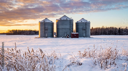 Three grain silos stand on a snow-covered farm field beneath a dramatic golden winter sunset sky