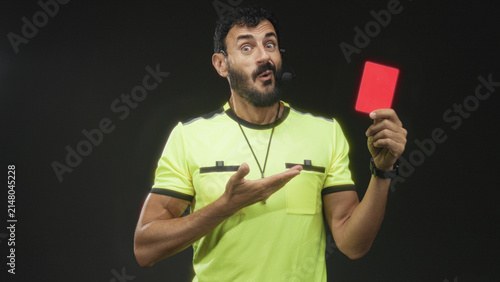 Fotografie Man referee in studio holds red card in raised hand and gestures with open palm, wearing whistle and headset; authority fairness discipline