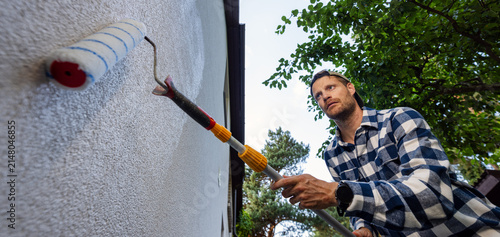 House painter applying protective coating on house exterior wall with roller. Facade protection against algae, moss and fungal