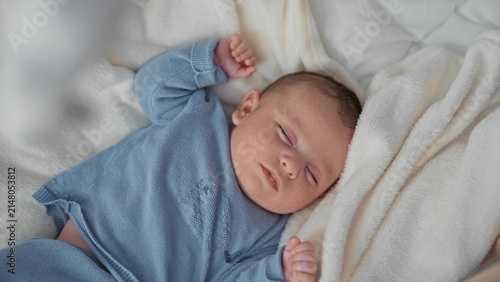 Cuadro en lienzo Boy sleeping with face visible on a bed in a nursery, wrapped in blue knit sweater and plush white blanket with arms relaxed; calm gentle rest