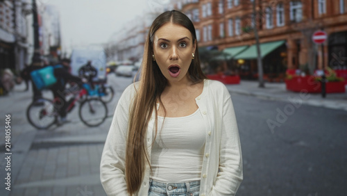 Obraz na plátne Young woman with open mouth gasping on a busy city street with delivery cyclists, cars and brick storefronts; surprise urgency