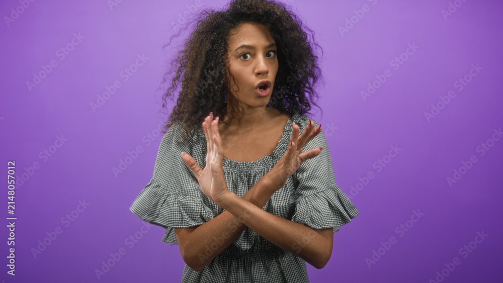 Obraz premium Curly haired woman with crossed hands signaling stop and bare hands visible in a purple studio; rejection refusal boundary.