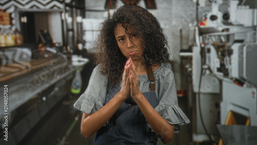 Photography Woman waitress with curly hair and apron, hands clasped pleading in a busy restaurant kitchen, pout expression and closed eyes; apology fatigue