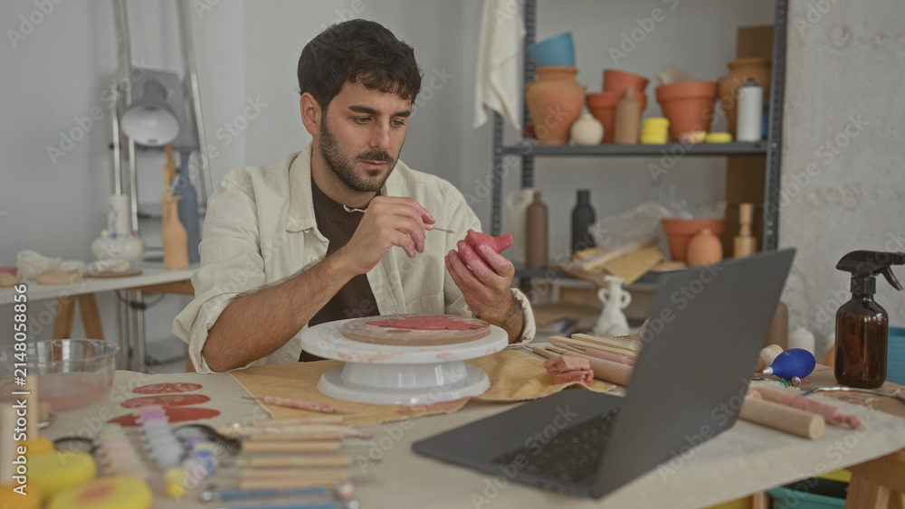 Fototapeta premium Man sculpts pink clay pot with hands on a spinning pottery wheel in studio, using loop tool and sponge while watching a laptop; concentration creativity.