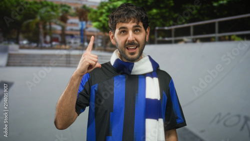 Fotografía Young bearded man in football shirt and scarf points finger upward on street in front of skate ramp; pride camaraderie