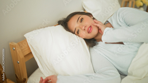Photography Woman lying in bed on pillow, hand to forehead covering eye, gentle smile while waking in a bedroom; serenity morning comfort