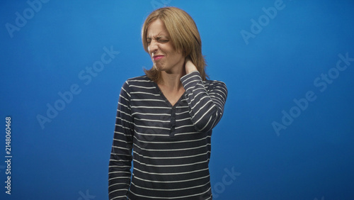 Fotografie Woman rubbing neck with right hand, wincing and holding her bare neck, striped top against blue backdrop in studio; discomfort recovery
