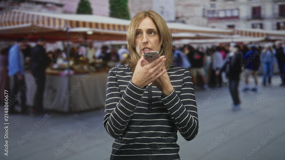 Fototapeta premium Woman holding smartphone to mouth speaking into speakerphone with expressive hand gesture among crowded outdoor market stalls; frustration urgency.