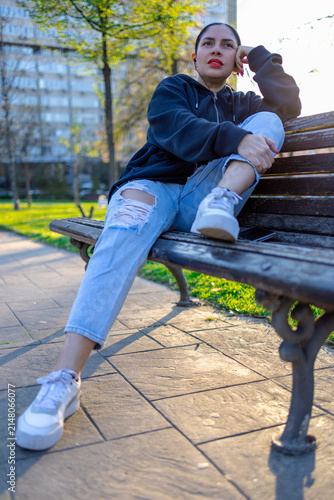 Young woman in a black hoodie, distressed light blue ripped jeans, and white sneakers relaxing on a park wooden bench during a sunny afternoon with trees and a building in the background.