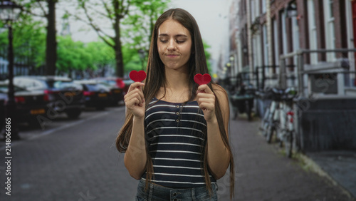 Fotografie Teen girl holds two red felt hearts between thumb and forefinger, eyes closed and lips pressed, standing on a city street with parked cars and bicycles; melancholy longing