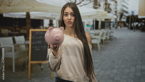 Photography Young woman holding pink piggybank forward with outstretched hand and sad expression on street cafe terrace; financial concern