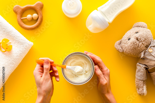 Make mixture for feed baby. Spoon with food in hand near toys and accessories on yellow background top view pattern