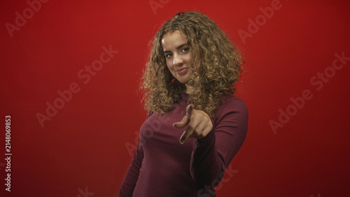 Canvas Print Young woman with curly hair beckons with her hand and gestures a finger point toward camera in studio with red backdrop; playful defiance