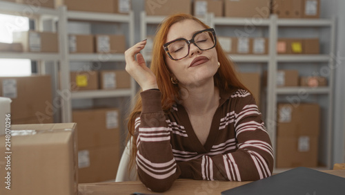 Quadro su tela Young woman with glasses resting head on hand at packing table with laptop and cardboard boxes on shelves in a building; tired monotony