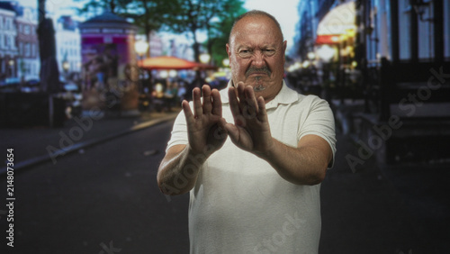 Fotografía Senior man with hands raised palms out pushing forward on city street, grimacing in discomfort; defensive disgust