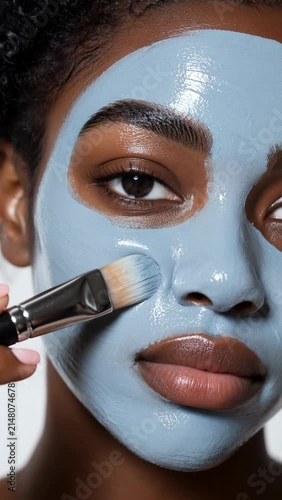 Close-up of a beautiful young Black woman applying a hydrating blue clay facial mask with a cosmetic brush for skin care and wellness.