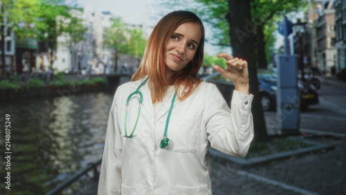 Canvastavla Redhead woman doctor in white coat with green stethoscope pinching fingers a small gap beside canal on a cobbled street; playful doubt