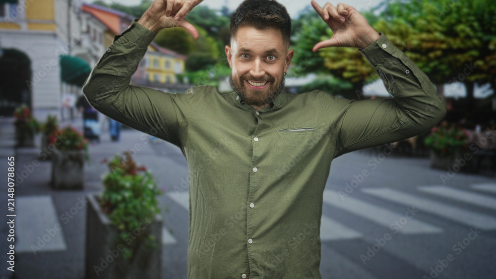 Fototapeta premium Man with beard using hands to frame face with thumb and forefinger on a pedestrian street, smiling at camera; spontaneous playfulness.